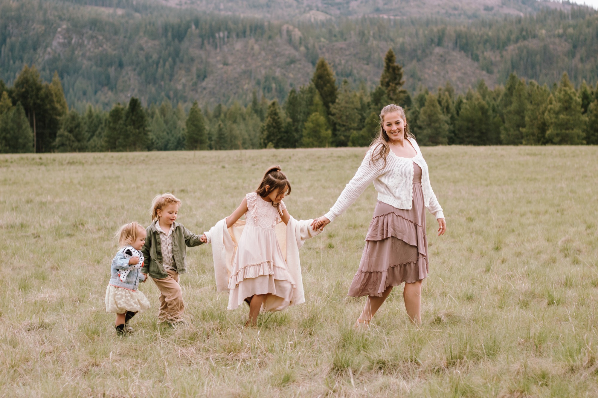 Mom smiling as she leads her three kids hand in hand through a golden North Idaho meadow with mountains behind them