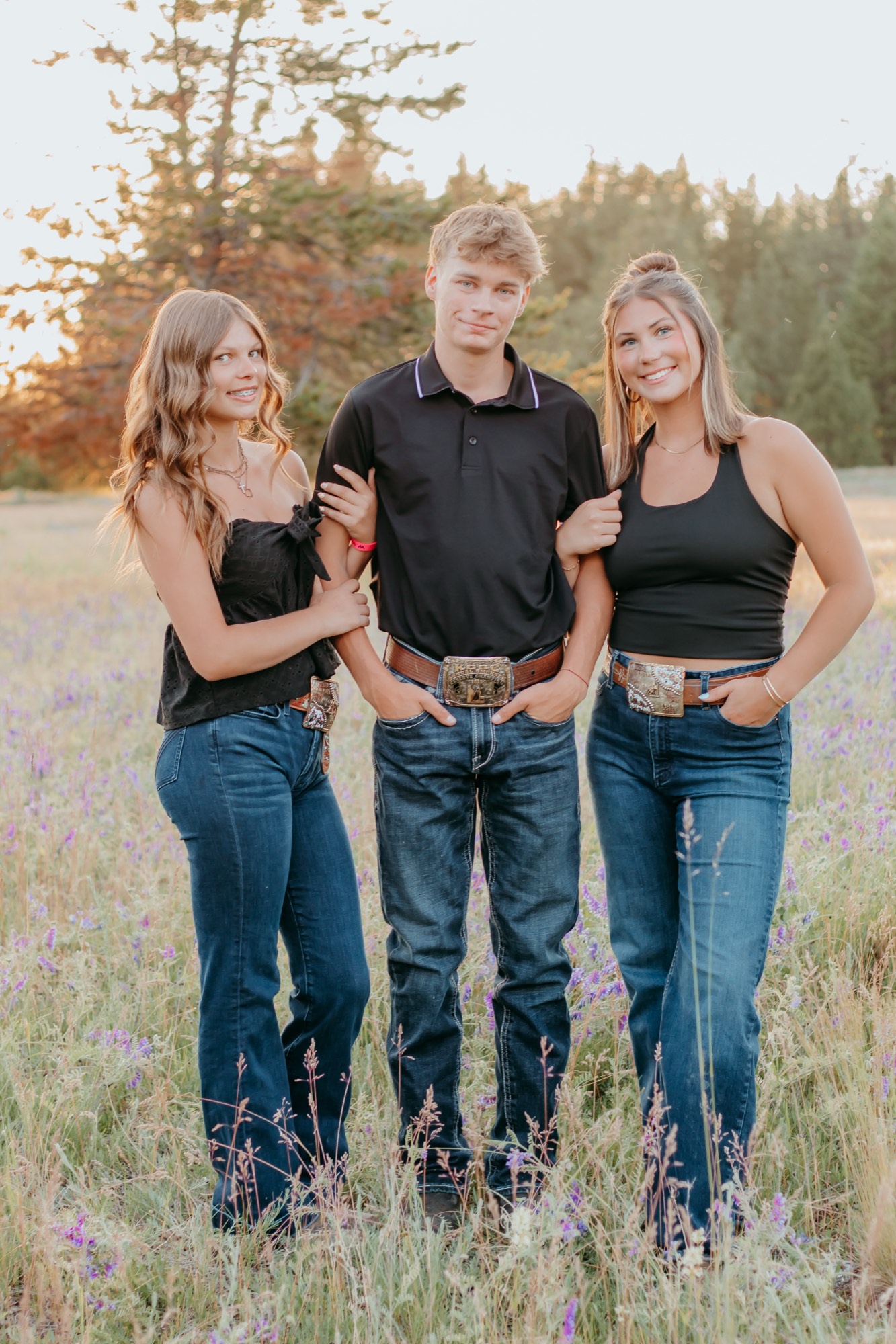 Three teens wearing rodeo championship belt buckles in a wildflower field at sunset near Post Falls Idaho